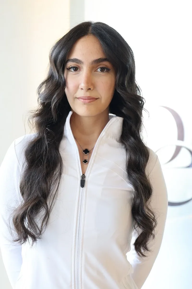 Woman with long dark wavy hair in a white zip jacket indoors