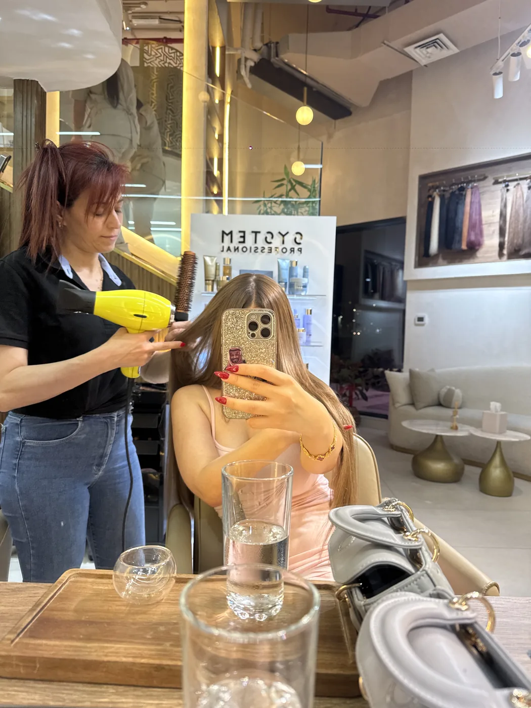 Stylist blow-dries a seated client's long hair with a round brush, mirror view