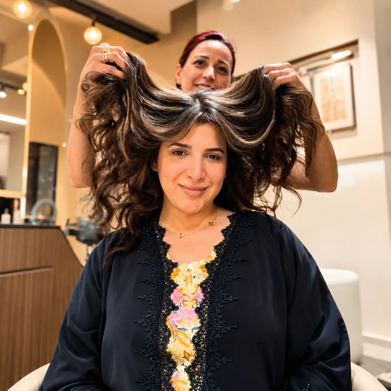 Stylist lifts a smiling client's long highlighted hair with both hands in salon