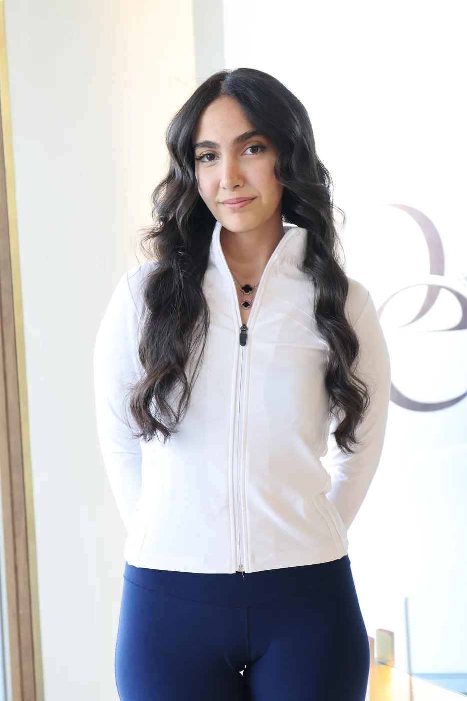 Young woman with long dark waves in a white jacket beside a frosted glass panel