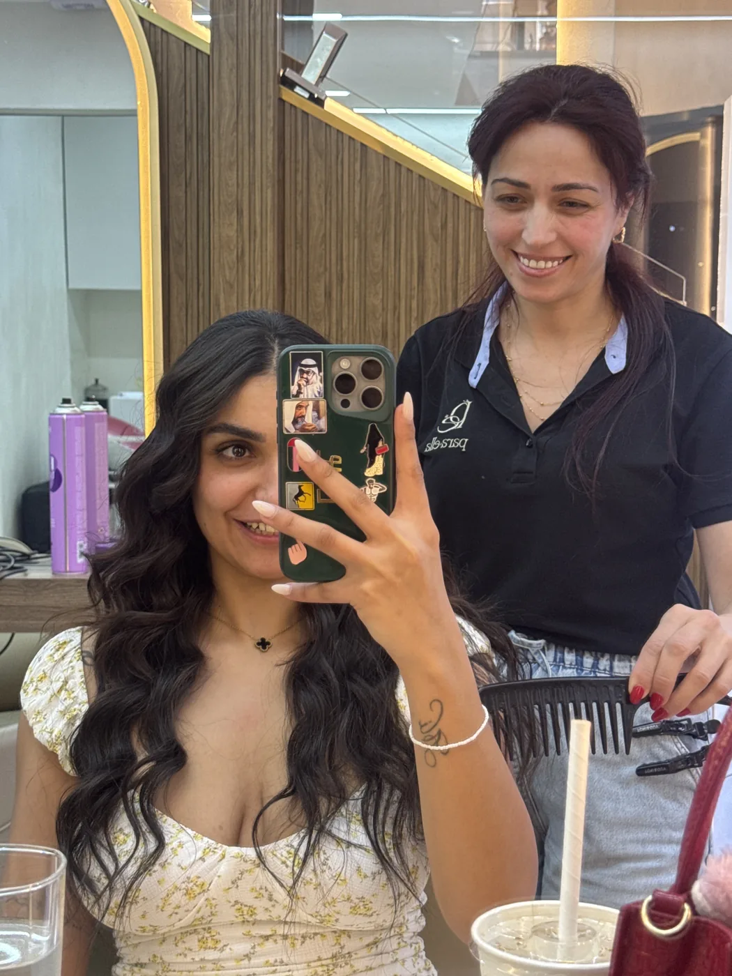 Mirror selfie of client with long dark waves, smiling stylist standing behind her