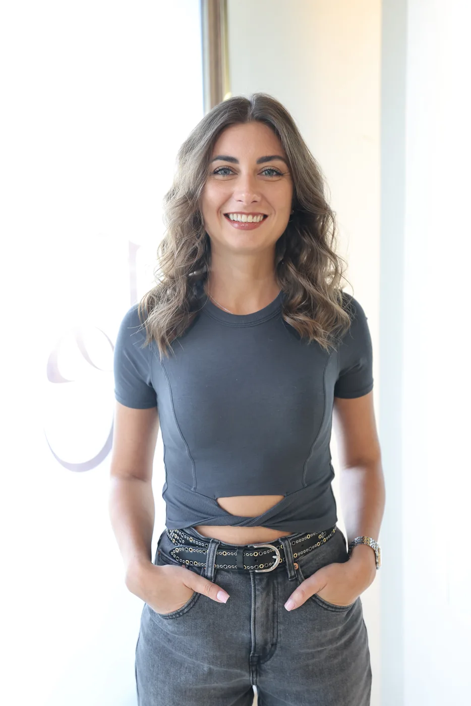 Woman with long wavy highlighted hair smiling, hands in jeans pockets, plain wall