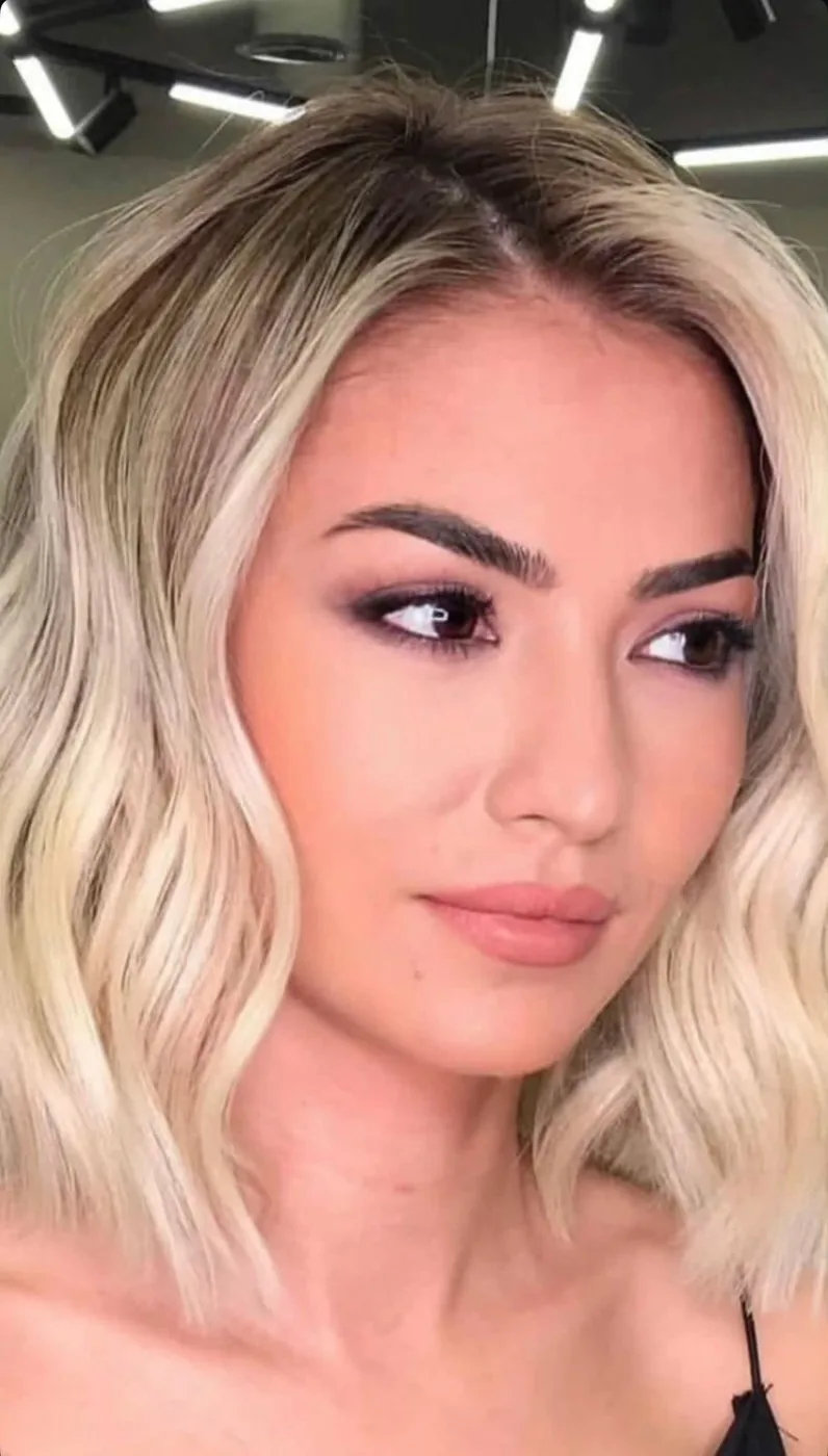 Woman with shoulder-length wavy blonde hair and full makeup under salon lights