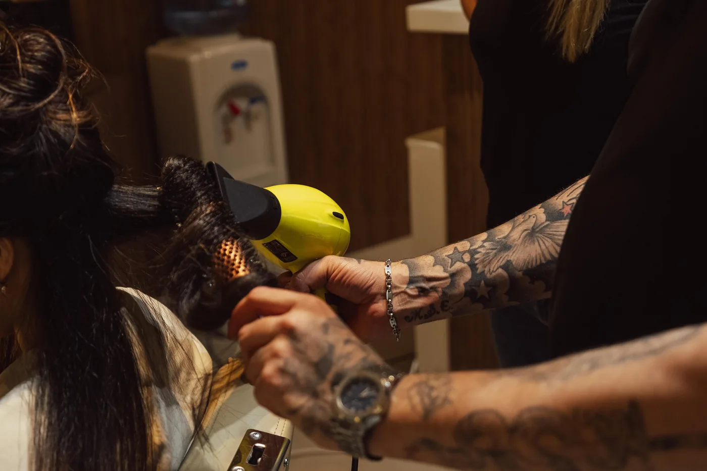 Stylist with tattooed forearms blow-drying a client's long dark hair using a round brush