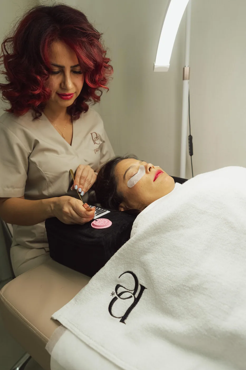 Technician in a beige uniform applying lash extensions to a reclining client