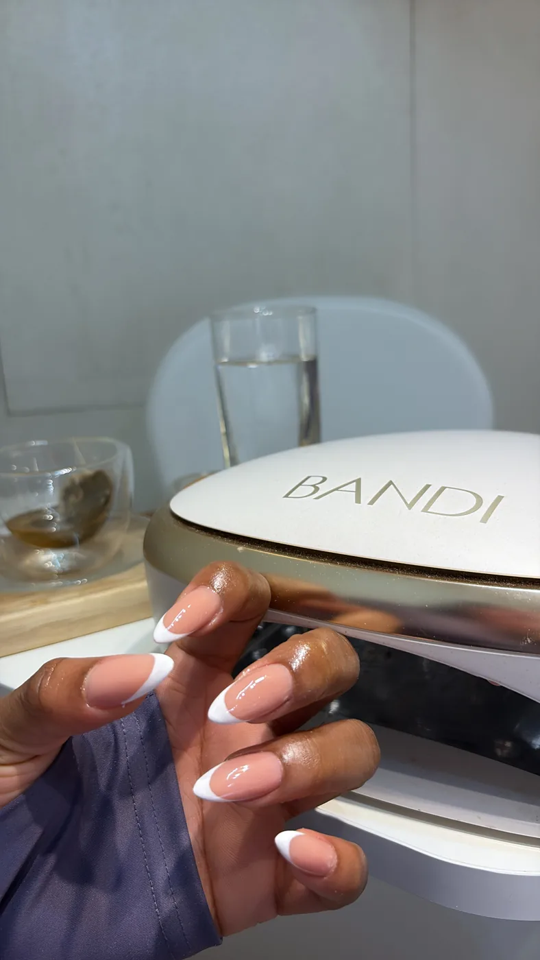Hand with long French manicure nails beside a white and gold BANDI nail lamp