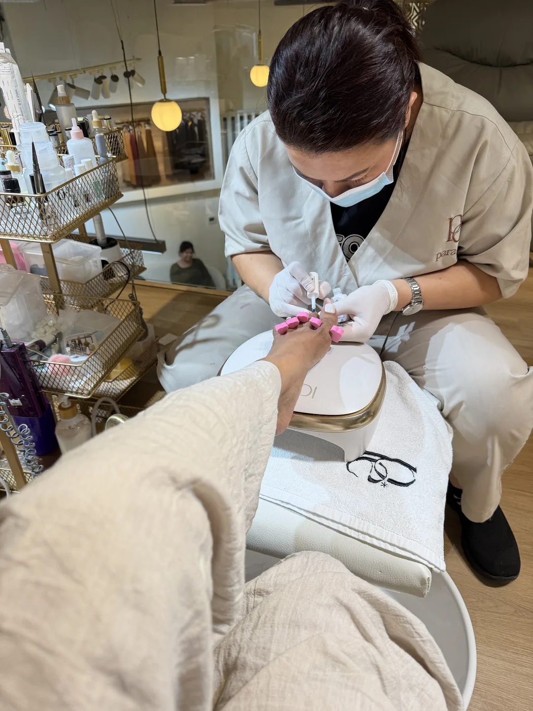 Masked technician in white gloves works on a client's bright pink toenails during a pedicure