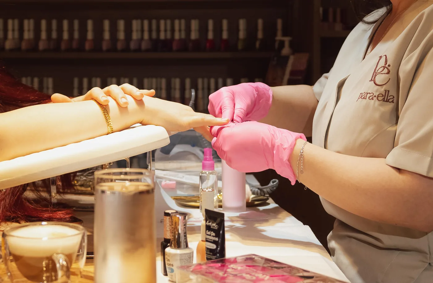 Nail technician in pink gloves works on a client's hand, uniform embroidered para·ella