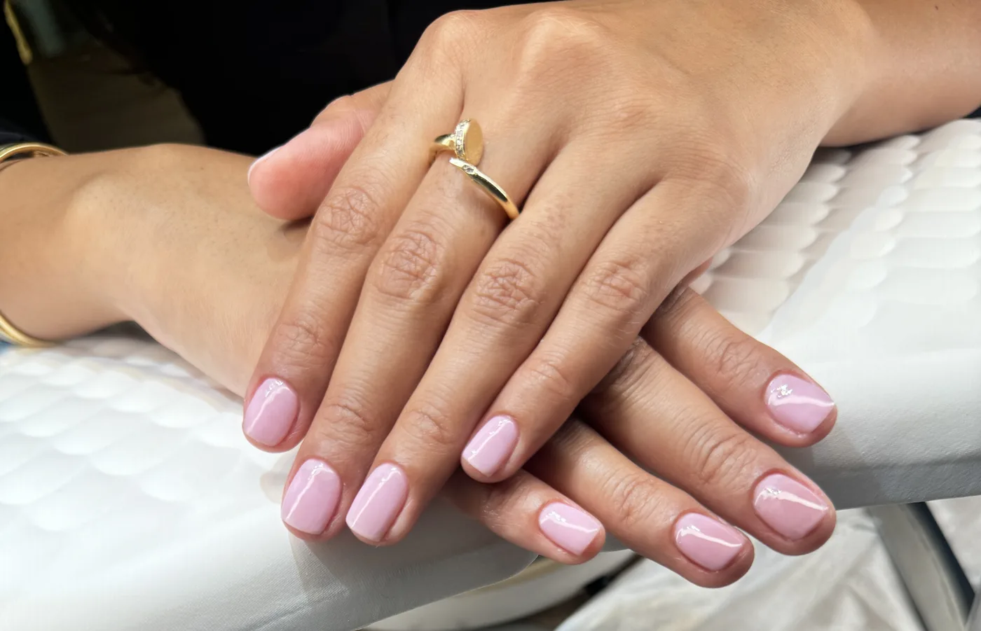 Two hands with glossy pale pink manicured nails resting on a white quilted cushion