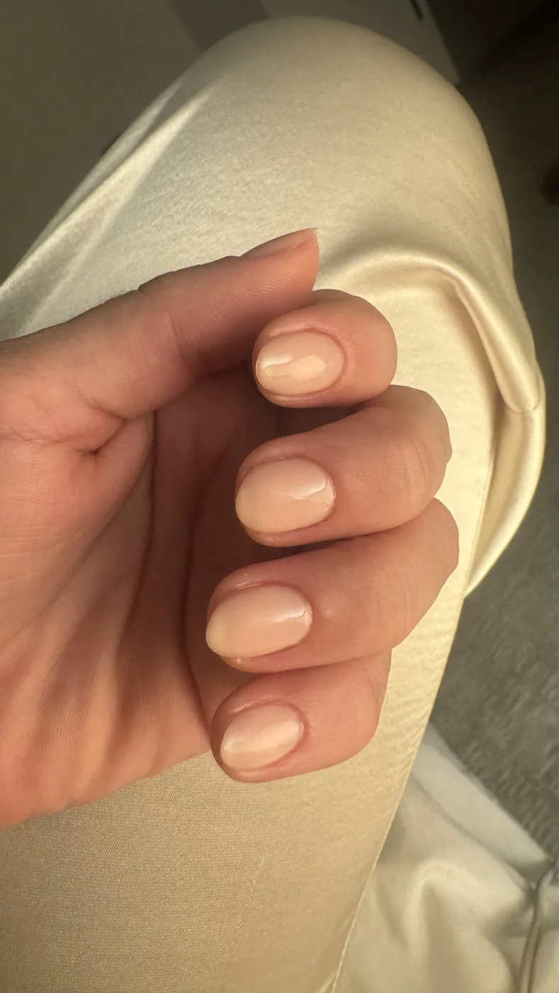 Hand with short rounded nails in sheer nude polish against cream satin fabric