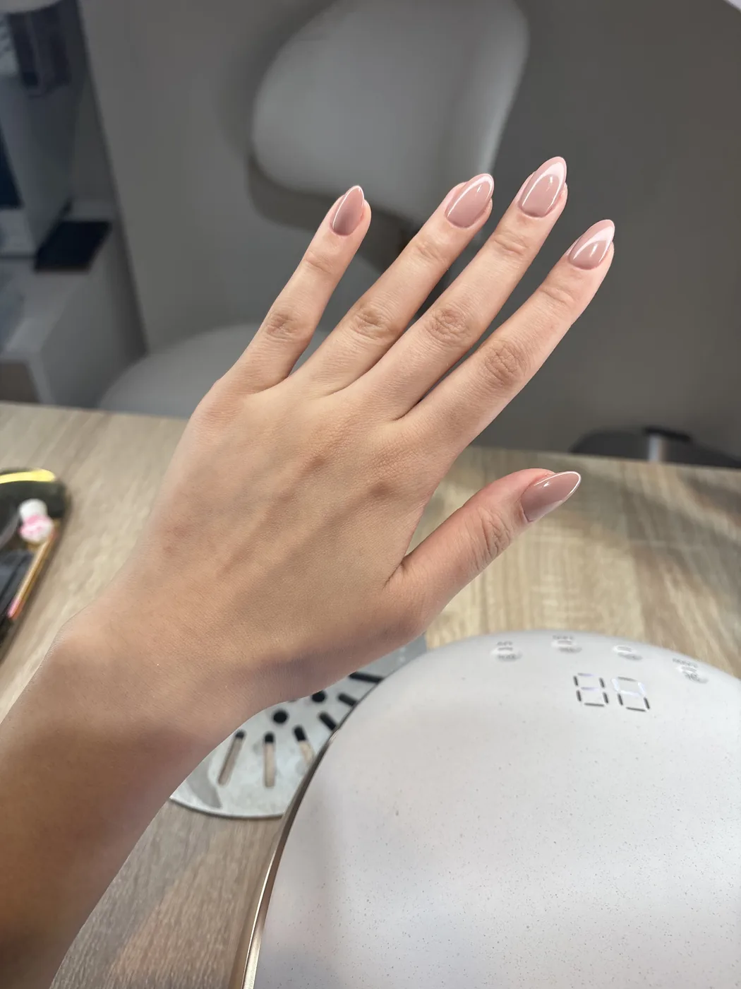 Hand with nude almond nails beside a white UV lamp on wooden salon table