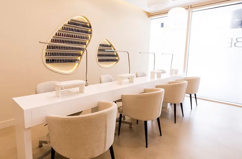 Manicure area with long white table, beige chairs and lit wall polish displays
